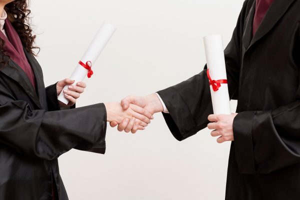 Δύο άτομα με ακαδημαϊκές ρόμπες ανταλλάσσουν χειραψία κρατώντας διπλώματα, σχετικό με τα μη κρατικά πανεπιστήμια.