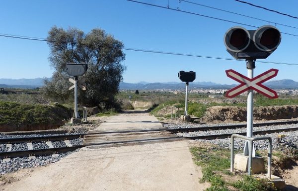 Σιδηροδρομική διάβαση με σήματα και δέντρο σε αγροτική περιοχή, θυμίζοντας τα παθήματα που δεν γίνονται μαθήματα.