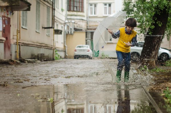 Αγόρι με ομπρέλα παίζει σε λακκούβες με νερό και λασποβροχές, απολαμβάνοντας τον καιρό.