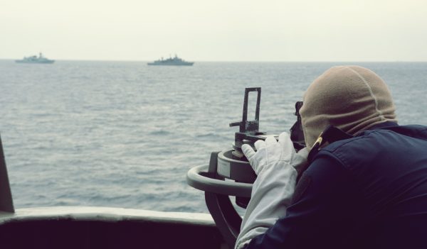 Άνθρωπος με γάντια χρησιμοποιεί ναυτικό όργανο για να παρατηρήσει πλοία στη θάλασσα, ενόψει του «ΠΑΡΟΝ» που κυκλοφορεί εκτάκτως.