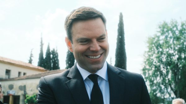 Ο Στέφανος Κασσελάκης χαμογελάει φορώντας κοστούμι σε εξωτερικό χώρο, με φόντο κτίριο και δέντρα.