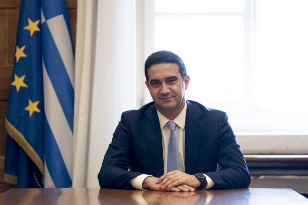 Ο Μιχάλης Κατρίνης κάθεται στο γραφείο του με φόντο τις ελληνικές και ευρωπαϊκές σημαίες, εστιάζοντας στην δικογραφία.