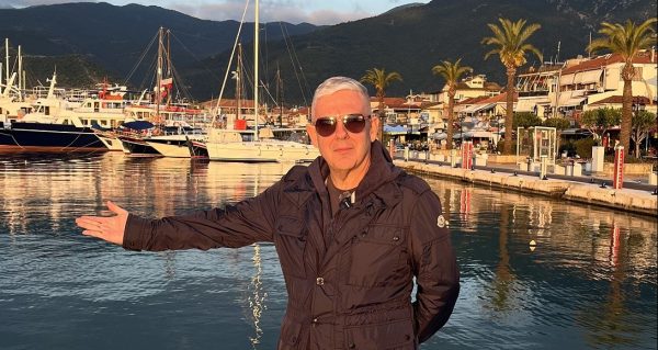 Ο Τάσος Δούσης ποζάρει στο λιμάνι της Λευκάδας, με τα ιστιοπλοϊκά και τα καταστήματα να διακρίνονται στο φόντο.