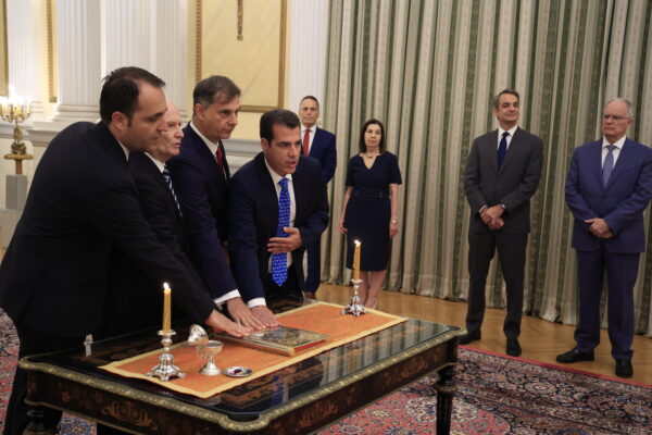 Νέα μέλη της κυβέρνησης ορκίζονται με τα χέρια πάνω σε ιερό βιβλίο, παρουσία του Κυριάκου Μητσοτάκη.