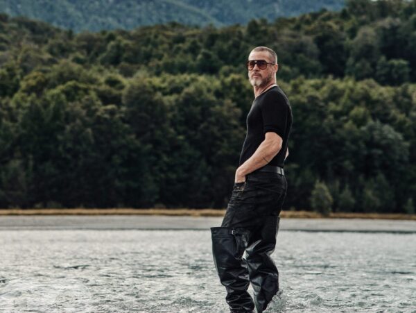 Ο Brad Pitt στέκεται σε νερά ποταμού με φόντο δάσος, φορώντας γυαλιά ηλίου και μπότες.