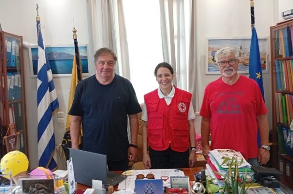 Τρεις άνδρες ποζάρουν για την αιμοδοσία του Ελληνικού Ερυθρού Σταυρού στην Κάσο, με τη σημαία της Ελλάδας και της ΕΕ στο παρασκήνιο.