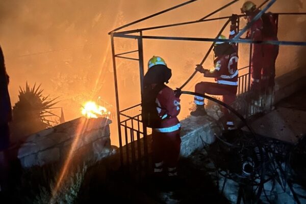 Εθελοντές του Ελληνικού Ερυθρού Σταυρού δίνουν μάχη με τις φλόγες, επιχειρώντας να σβήσουν τη φωτιά.