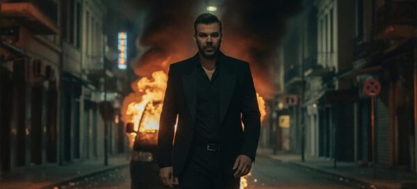 Ο Γιώργος Σαμπάνης περπατάει σε δρόμο με φλεγόμενο αυτοκίνητο στο βίντεο κλιπ του «Προς Το Παρόν».