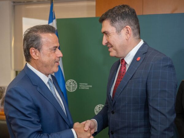 Δύο άνδρες ανταλλάσσουν χειραψία, με φόντο την ελληνική σημαία και το λογότυπο της Coca-Cola Τρία Έψιλον για την αποκατάσταση πυρόπληκτων περιοχών.