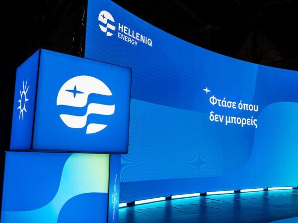 Λογότυπο της HELLENiQ ENERGY και το σλόγκαν «Φτάσε όπου δεν μπορείς» σε σκηνικό εκδήλωσης.