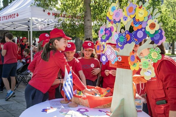 Παιδιά με κόκκινες μπλούζες και καπέλα του Ε.Ε.Σ. στήνουν περίπτερο με δέντρο ευχών και υποστηρίζουν παιδιά που χρειάζονται στήριξη.