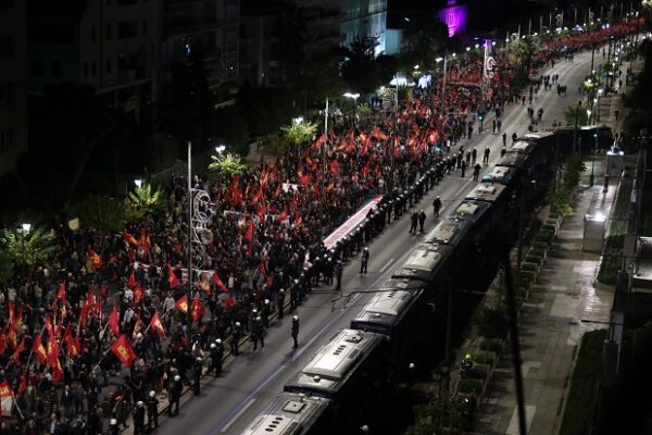 Πλήθος διαδηλωτών με κόκκινες σημαίες και αστυνομικές δυνάμεις σε πορεία κατά τη διάρκεια νυχτερινής συγκέντρωσης.