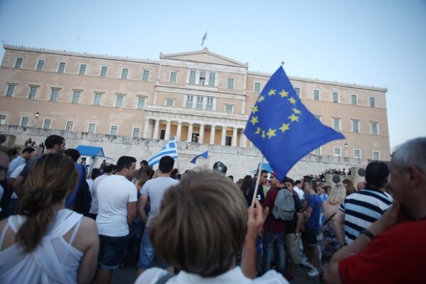 Συγκέντρωση πολιτών μπροστά από τη Βουλή κρατώντας σημαίες Ελλάδας και Ευρωπαϊκής Ένωσης σε κρίσιμη κοινωνία.