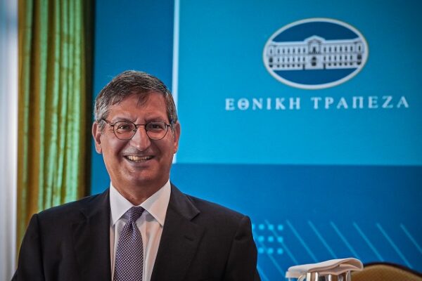 Ο κ. Μυλωνάς χαμογελά μπροστά από το λογότυπο της Εθνικής Τράπεζας, στο πλαίσιο των ιστοριών τρέλας.