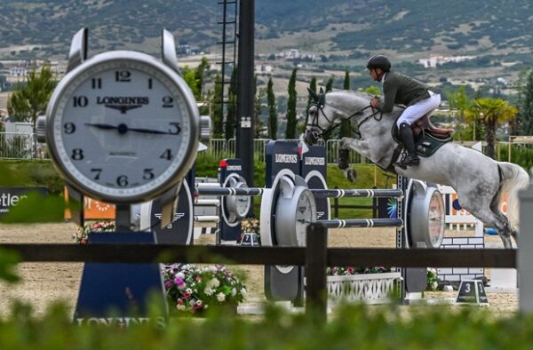 Αναβάτης με άλογο σε άλμα κατά τη διάρκεια του Ippos Spring Tour 2026, με μεγάλο ρολόι Longines στο προσκήνιο.