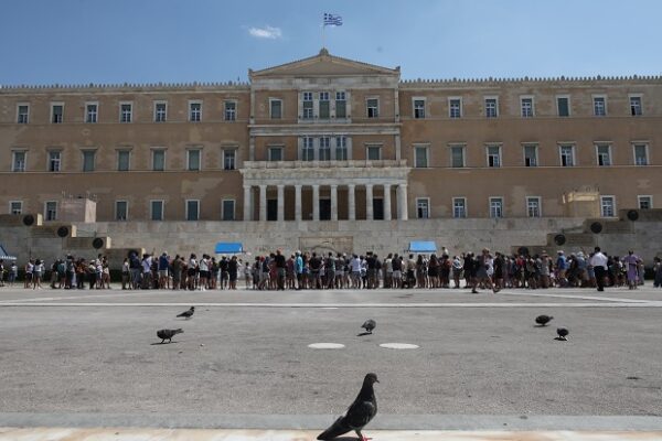 Πλήθος κόσμου συγκεντρώνεται μπροστά από το κτίριο της Βουλής των Ελλήνων, με περιστέρια να περπατούν στην πλατεία.