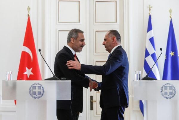 Δύο άνδρες χαιρετούνται μπροστά από ελληνικές και τουρκικές σημαίες, σχολιάζοντας τα ήρεμα νερά.