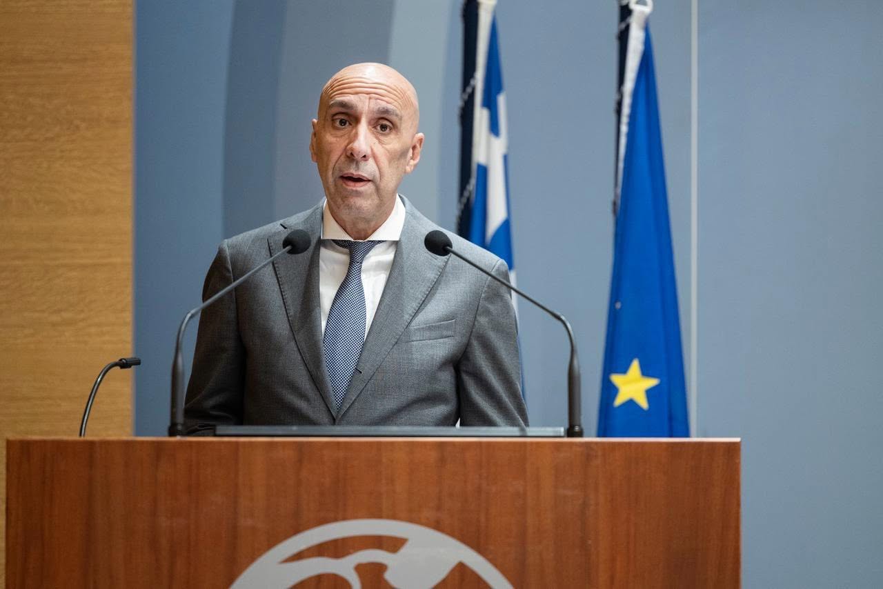 Ο Γιάννης Μπρατάκος μιλάει από το βήμα, με τις σημαίες της Ελλάδας και της ΕΕ να διακρίνονται στο βάθος, αναφορικά με τις εξαγγελίες στη ΔΕΘ.
