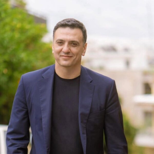 Ο Β. Κικίλιας στην Κύπρο για την 9η Τριμερή Συνάντηση για τη Ναυτιλία, με φόντο πράσινο και κτίρια.