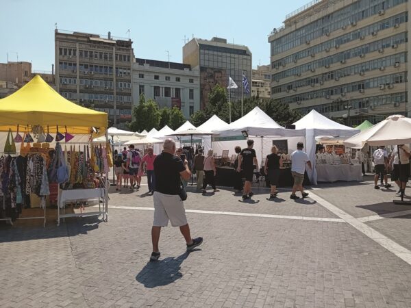 Υπαίθρια αγορά με πάγκους και κόσμο σε πλακόστρωτο, με κτίρια στο φόντο.